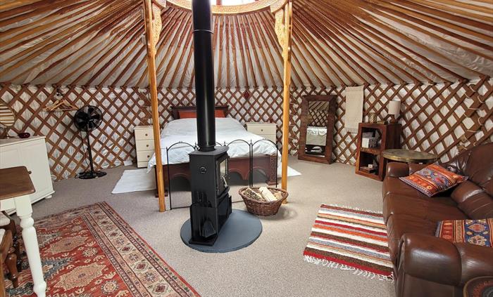 Yurt interior is round, with many carved wood rafters radiating from a carved wood central roof light. Wood bed to rear, brown leather sofa to right,